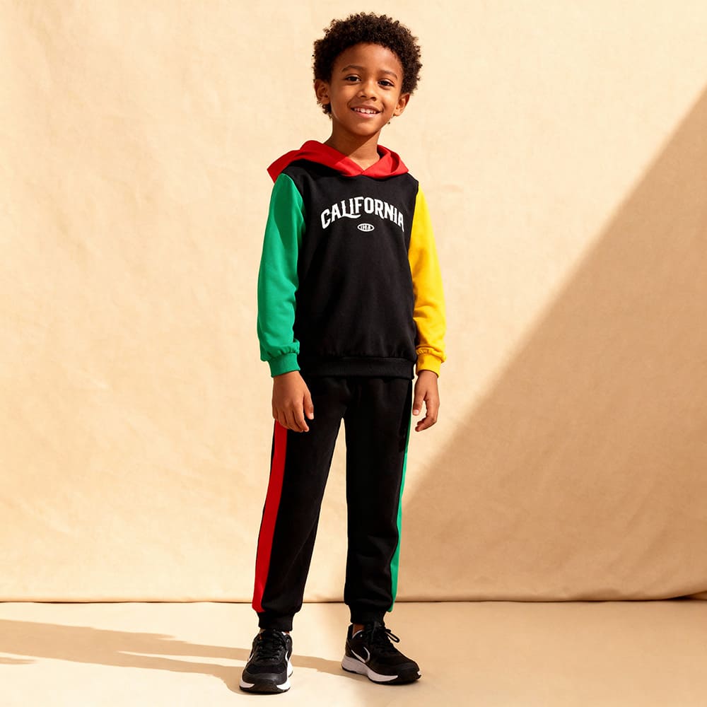 california-hoodie-boy-model-styles-by-syl-2 Black colorblock hoodie with “CALIFORNIA” graphic, red hood, yellow & green sleeves + matching black joggers with striped sides — sporty, bold outfit for boys.