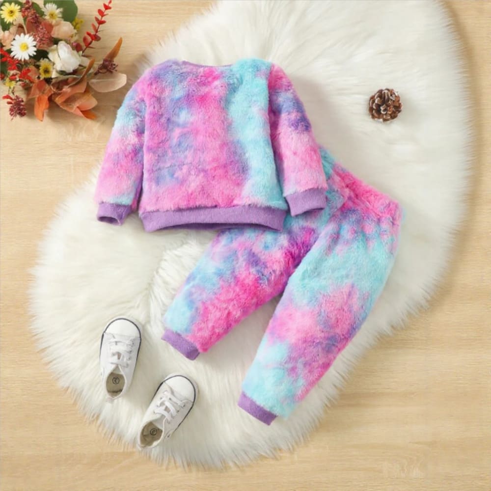 A vibrantly colored toddler girls' fuzzy loungewear set in tie-dye shades of pink, purple, and blue. The set includes a long-sleeve crewneck pullover top and matching jogger pants with elastic cuffs, laid flat on a white fluffy rug.
